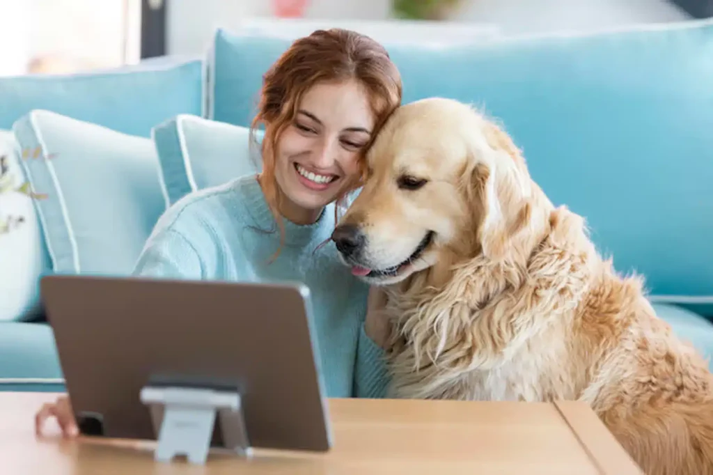 A smiling woman and a golden retriever sit close together on a couch, looking at a tablet on a coffee table in a cozy living room with blue decor.