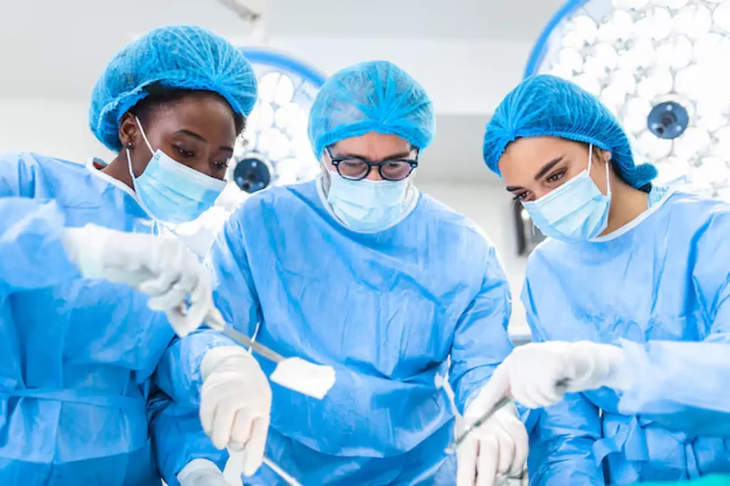 Three surgeons in blue scrubs, masks, and hair covers are focused on performing surgery under bright operating room lights, working together with surgical instruments.